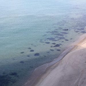 Aerial view of Mangsang Beach reveals the transparent East Sea waters meeting the sandy beach, with visible sand patterns and seaweed beneath the gentle waves.