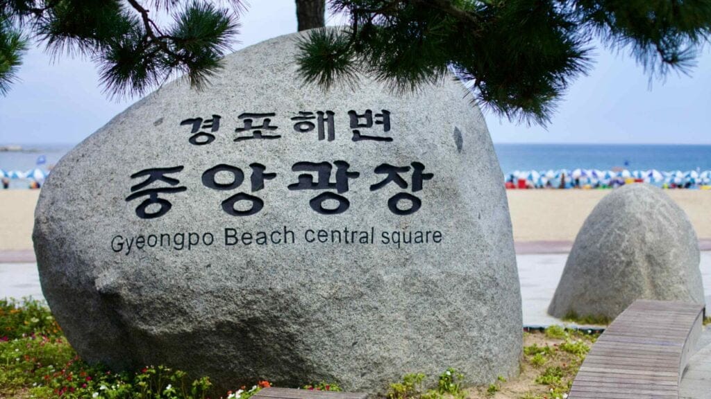 A stone marker inscribed with "Gyeongpo Beach Central Square" in both Korean and English, set against the sandy beach and the East Sea.