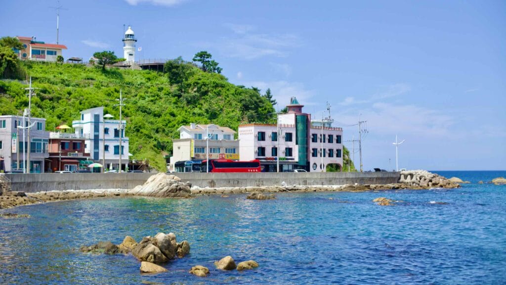 A picturesque seaside road just north of Jumunjin Port, nestled under a green hill with a quaint lighthouse atop, flanked by Minbak hotels beside the azure East Sea, with rocks dotting the waters.