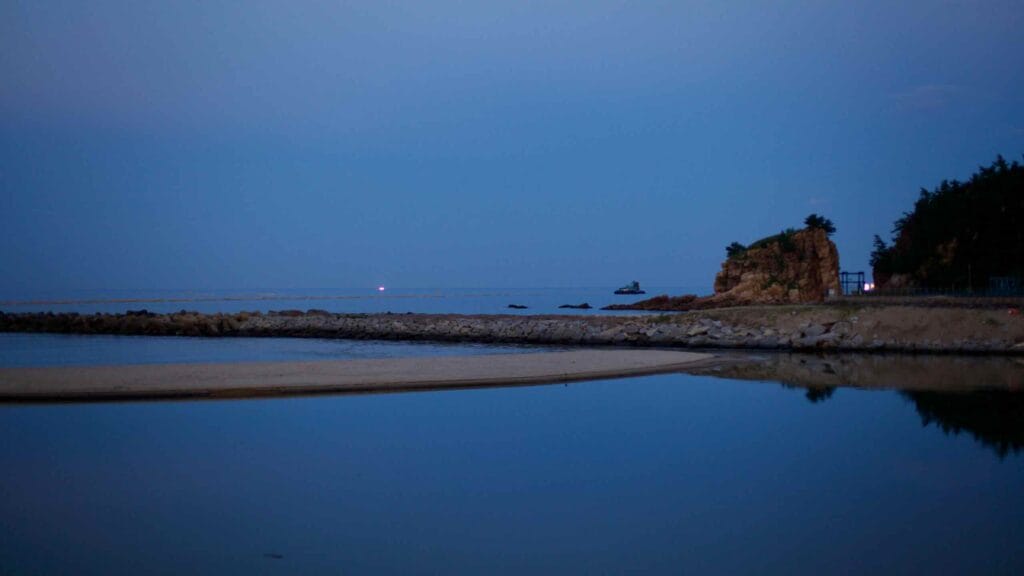 The early evening view of Anyinjin Port reveals calm waters of the East Sea, with light lingering on the horizon. Sandy banks and a small rocky outcrop mark the coastline, where sea and sky seem to meld into one serene landscape.