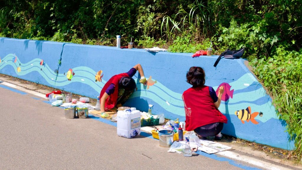 Along a road near Gajin Port, two painters bring life to a retaining wall, creating a vibrant mural.
