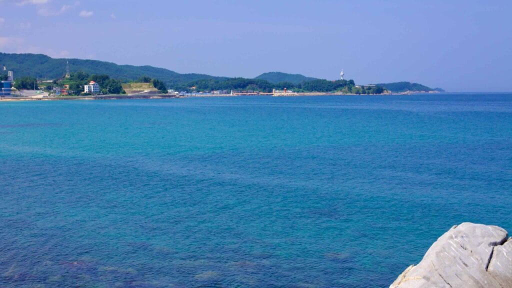 Overlooking the azure waters near Chodo Beach, the view captures green hills and a white lighthouse standing in the distance.