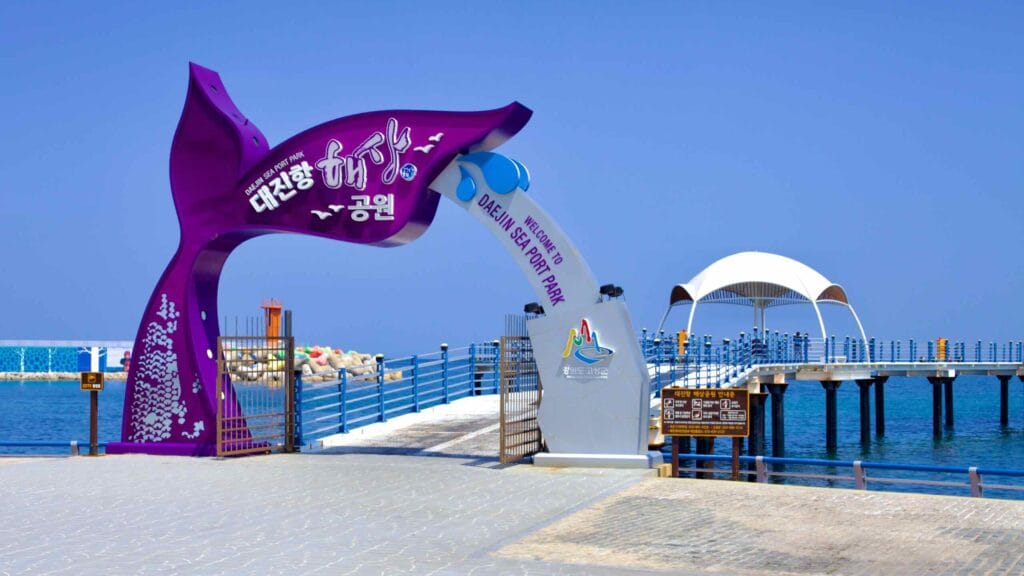 The entrance to Daejin Sea Port Park is marked by a purple whale tail arching over the walkway, leading visitors over water to a lookout platform.