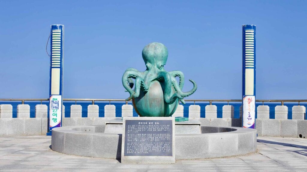 A whimsical octopus sculpture, perched atop a ball near the East Sea at Mukho Port, stands out against the backdrop of a clear blue sky, adding a playful touch to the coastal landscape.