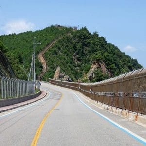 A coastal road near Geojin Port winds 53 meters up through a mountain pass, flanked by a military fence and the rocky coast of the East Sea.
