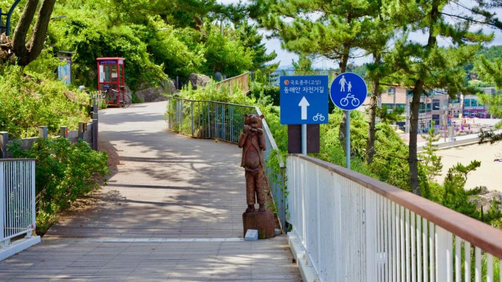 A walking and biking path winds towards the Chuam Chotdaebawi Certification Center nestled along a rocky ridge with Chuam Beach below and green hillsides around.
