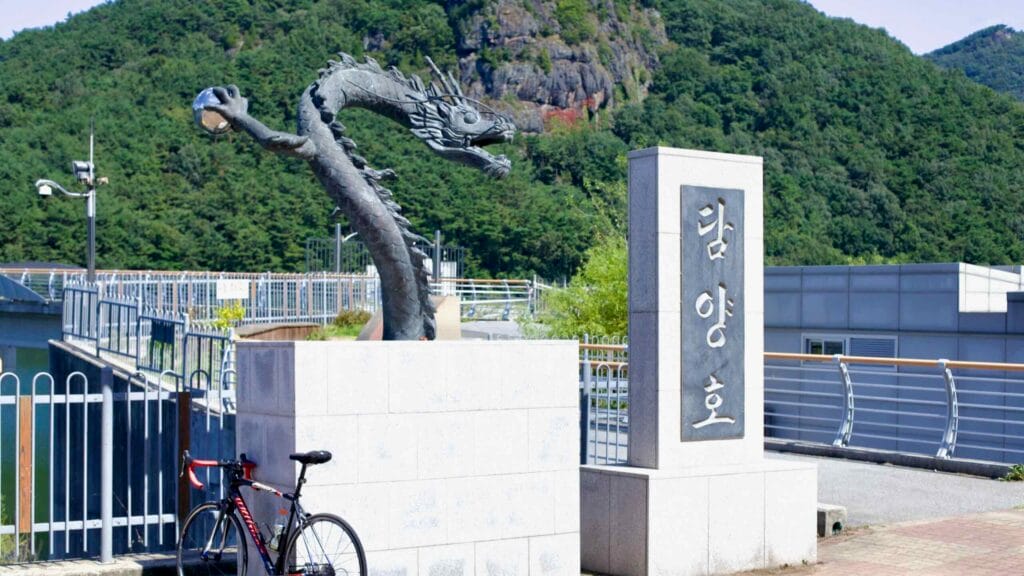 Road bike leans against a dragon statue at Damyang Lake, backed by forested hills.