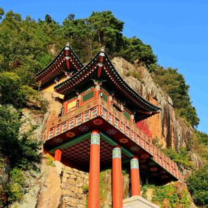 The golden light of sunset bathes Saseongam Hermitage, highlighting its dramatic cliffside architecture and the statues of guardian deities.