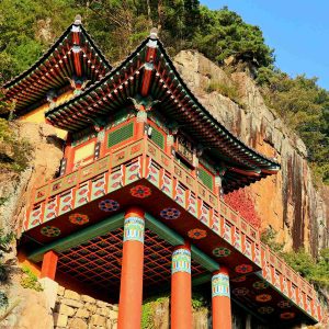 The golden light of sunset bathes Saseongam Hermitage, highlighting its dramatic cliffside architecture and the statues of guardian deities.