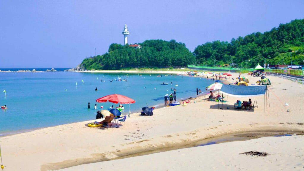 A southward view of Machajin Beach reveals beachgoers and the Daejin Lighthouse perched on a hill in the background.