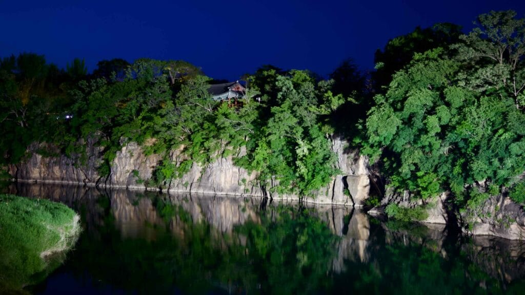 Jukseoru Pavilion illuminated at night, perched on a rocky clifftop, reflecting in the calm waters of the Osip Stream.