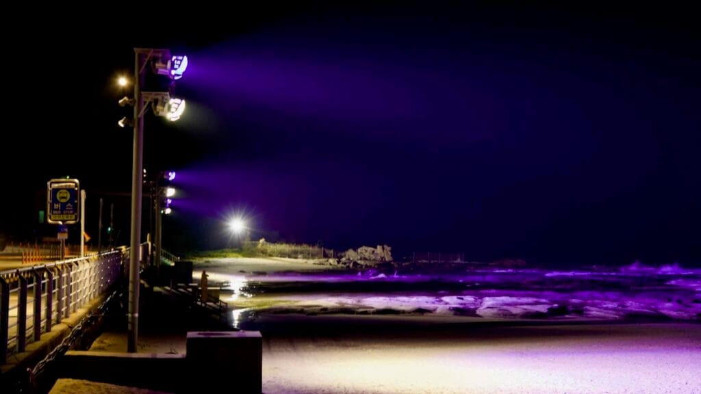 Purple and white lights illuminate Ayajin Beach at night, casting reflections over the sand and rocky shoreline.