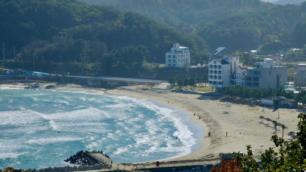 A view of Ingu Beach with its sandy shores and gentle waves, surrounded by lush green hills and nearby modern buildings, offering a serene coastal atmosphere.