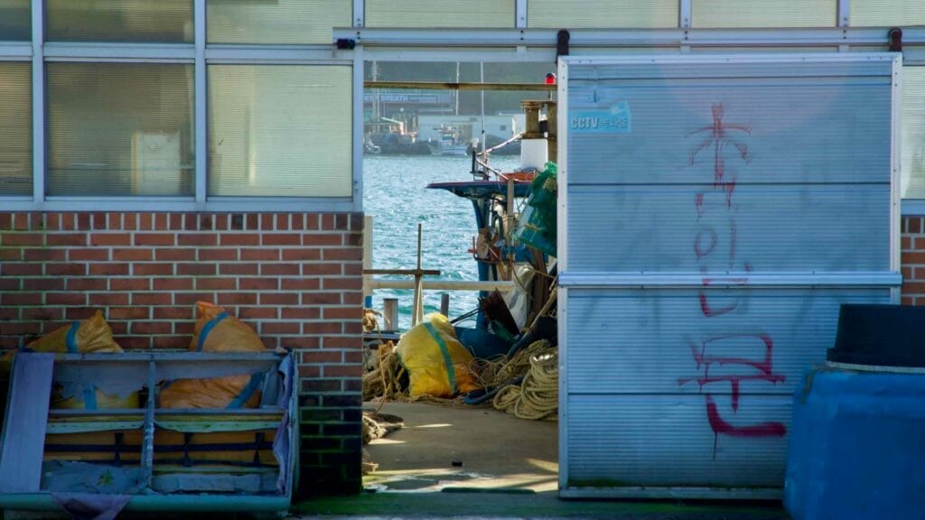 A peek through a doorway reveals the bustling docks of Namae Port, with fishing equipment, boats, and the glistening waters of the East Sea in the background.