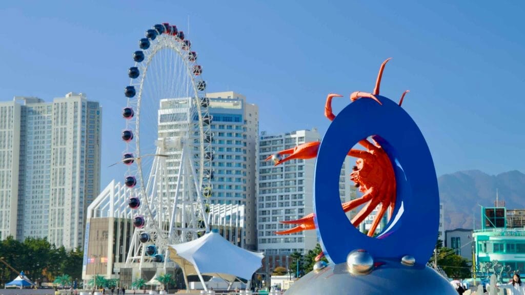 The Sokcho Eye Ferris Wheel and the vivid crab sculpture stand prominently against modern skyscrapers and the backdrop of Sokcho Beach, blending art with urban life.