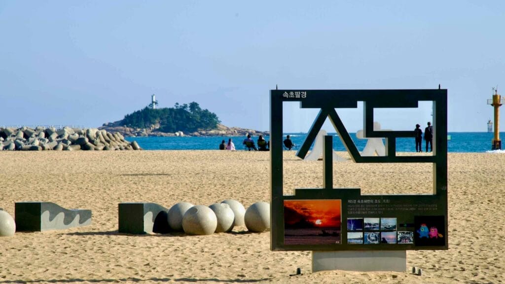 A scenic display at Sokcho Beach featuring a sign spelling out "Jodo Island," with the island's lighthouse and rocky terrain visible in the background.