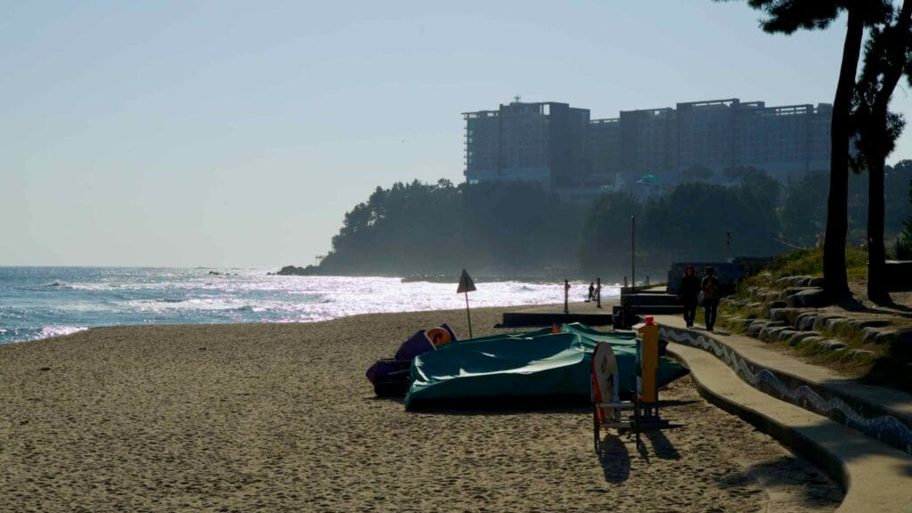 A serene view of Sokcho Beach during sunset, with waves shimmering in the light and the Lotte Resort perched on a forested hill in the background.
