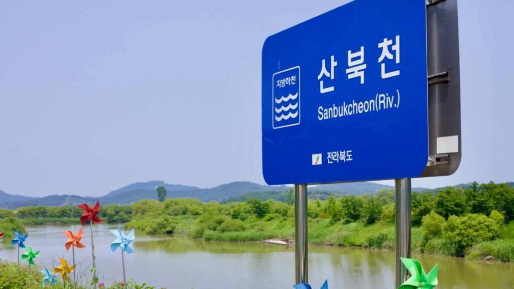 A blue sign marks the Sanbukcheon River, a tributary of the Geum River, with colorful pinwheels