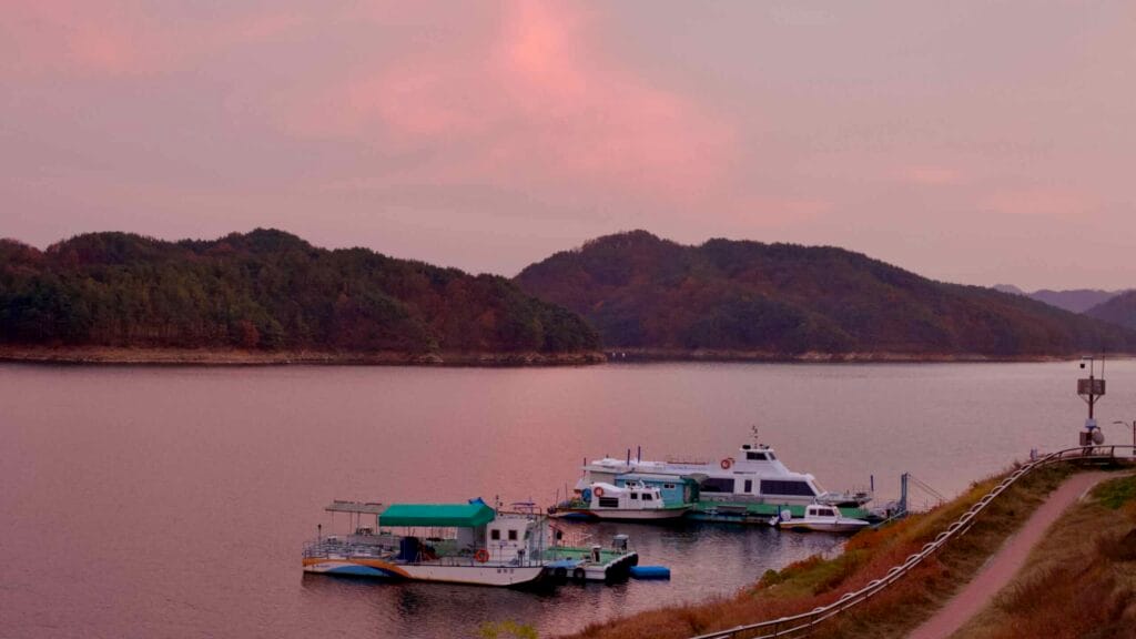 Boats docked along the calm waters of Daecheong Lake at sunset, with rolling forested hills in the background.
