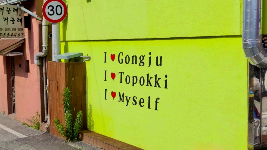 A bright yellow mural in downtown Gongju with playful text reading "I ♥ Gongju, I ♥ Topokki, I ♥ Myself."