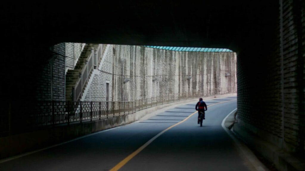 A cyclist rides through a dimly lit underpass along a curved road, emerging into daylight.