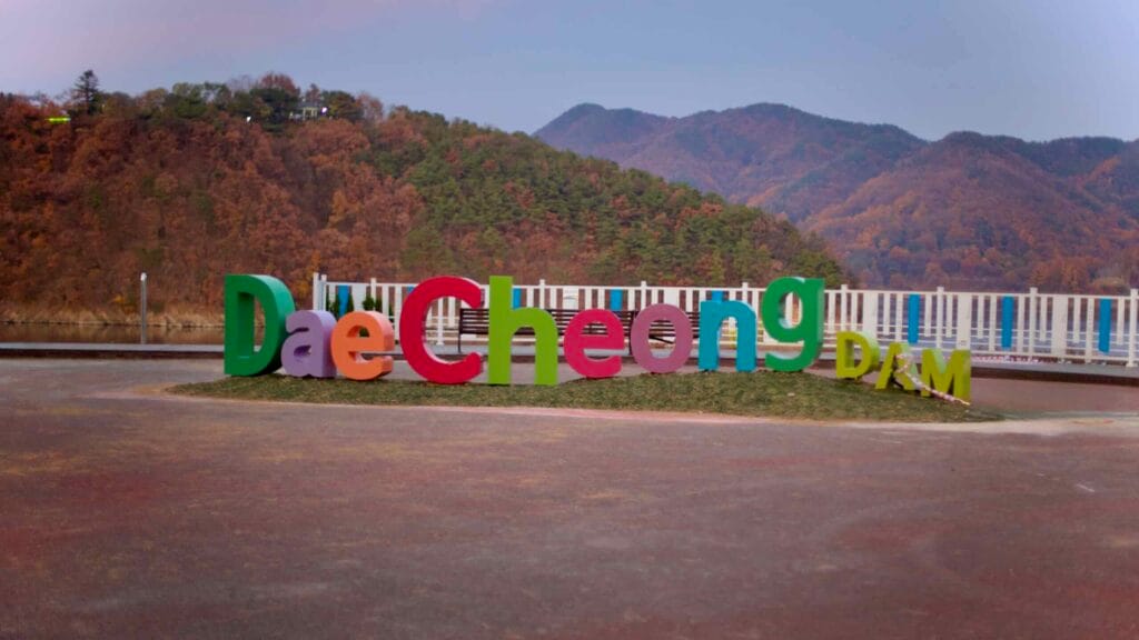 A Daecheong Dam sign stands against an autumn mountain backdrop at sunset.
