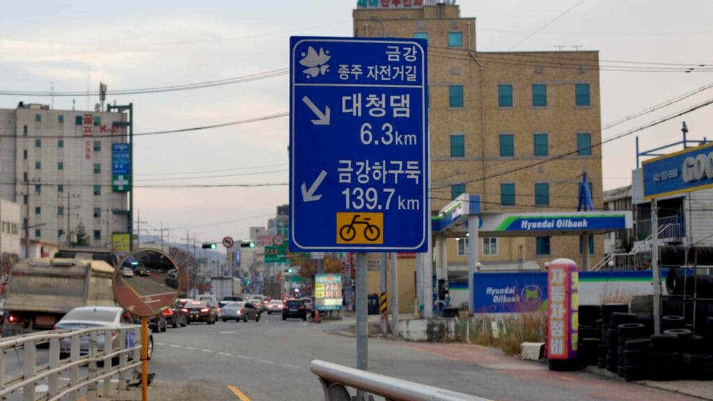A blue road sign along the Geum River cycling route provides directions to Daecheong Dam.