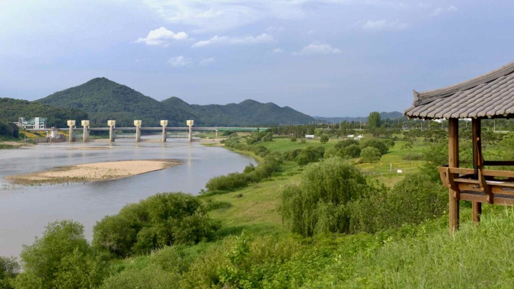 A view of the Geum River with Gongju Weir in the distance, surrounded by greenery and rolling hills.