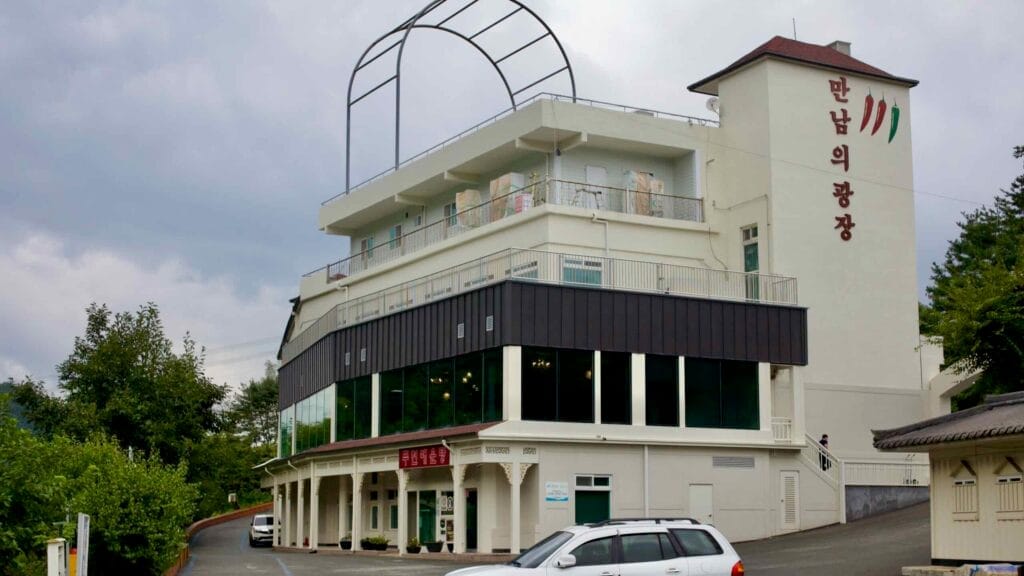The Mannameui Gwangjang building, a local establishment with a restaurant and event hall, stands along a rural road. Its modern design contrasts with the surrounding greenery.