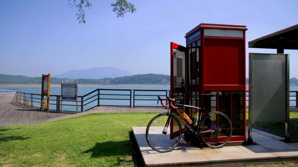A road bike leans against the Baealdo Waterfront Park certification booth, where cyclists stamp their bike passports. The booth sits beside a scenic boardwalk overlooking the Seomjin River, marking the start of the Seomjingang Bike Path.