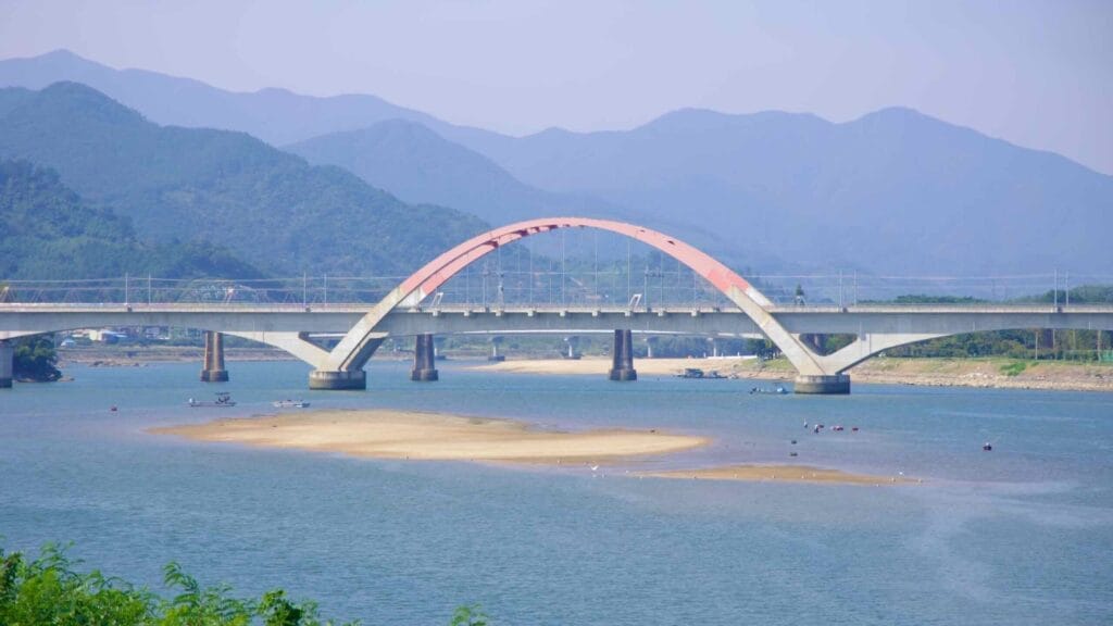 A modern railroad bridge with a distinctive arch design crosses the Seomjin River, with sandbanks visible below and mountains forming a scenic backdrop.