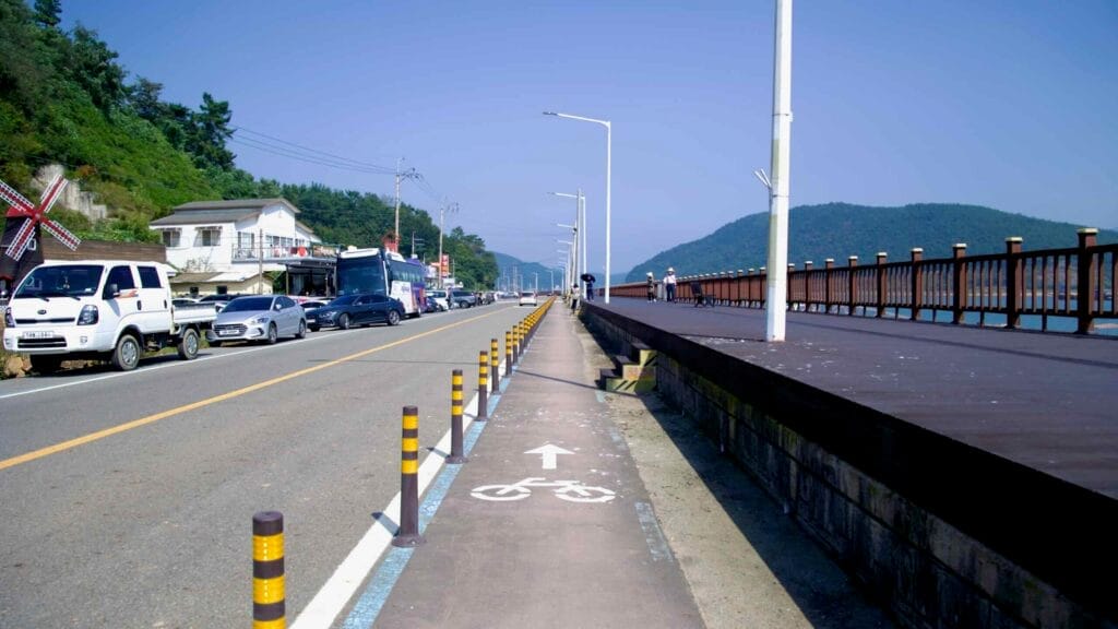 A designated bike lane runs parallel to a riverside boardwalk along the Seomjingang Bike Path. Cyclists and pedestrians enjoy scenic views while passing roadside shops and parked vehicles.