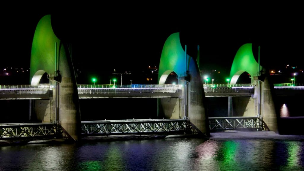 The Seungchon Weir on the Yeongsan River is illuminated with green lighting, casting vibrant reflections across the river at night.
