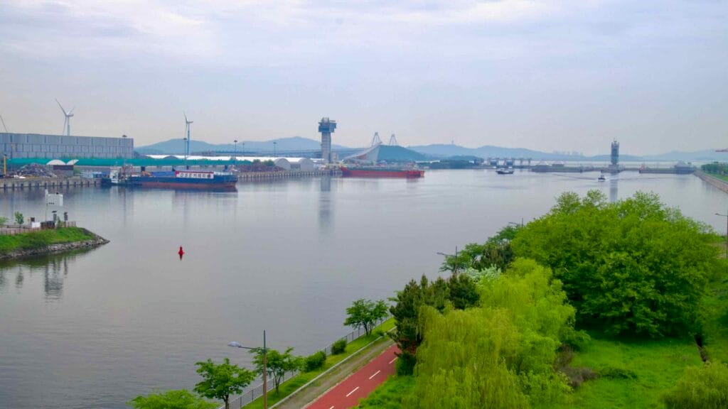 A panoramic view of the Ara Waterway and Incheon Port near the West Sea Lock, framed by lush greenery and a red pedestrian lane running parallel to the industrial waterfront.