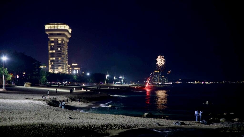 Fireworks and city lights illuminate Gyeongpodae Beach as beachgoers gather on the sand.