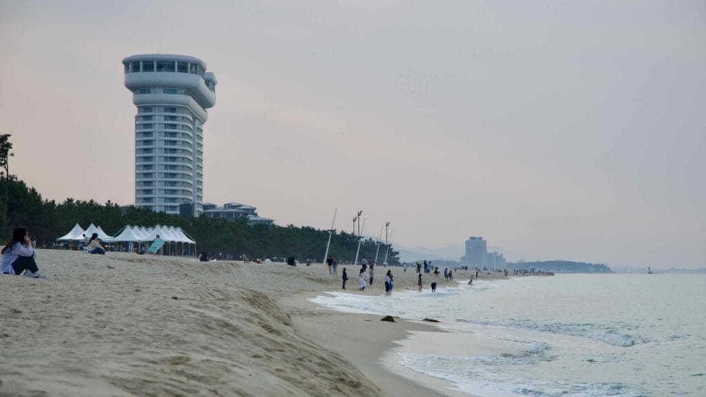 Hotel tower overlooking Gyeongpodae Beach, where visitors walk the waterline and tents dot the shore.