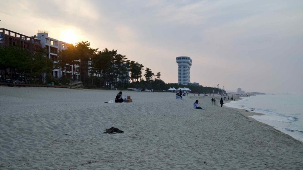 Evening at Gyeongpodae Beach: sun setting, beachgoers resting, and the round tower rising.