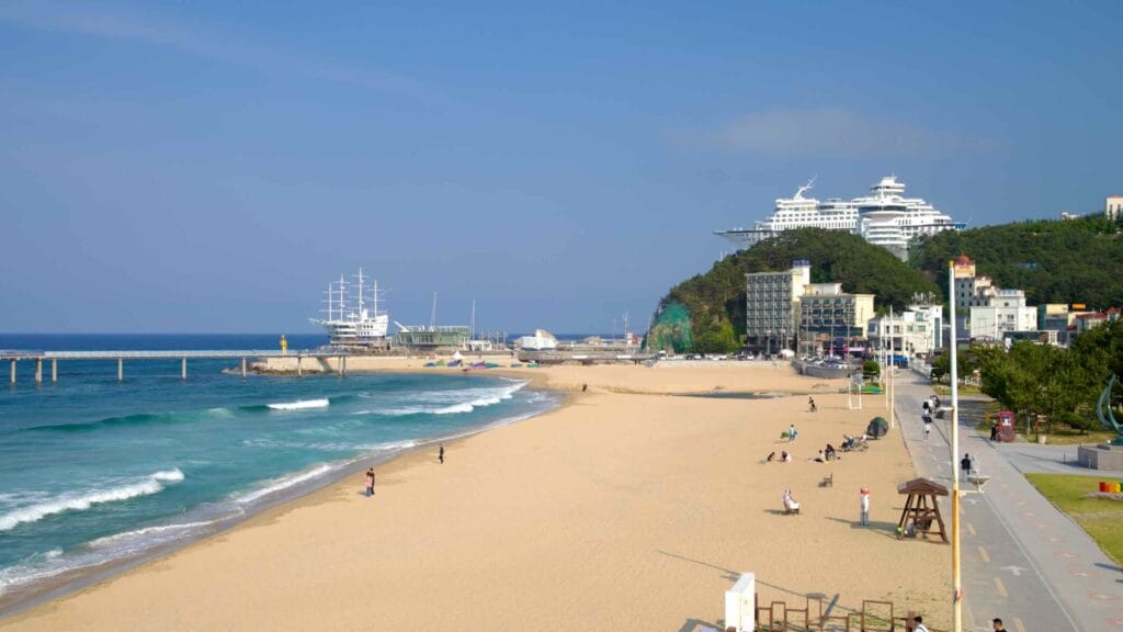 A wide view of Jeongdongjin Beach captures gentle waves, a long pier, and the cruise ship-shaped hotel rising above the coastline under a clear sky.