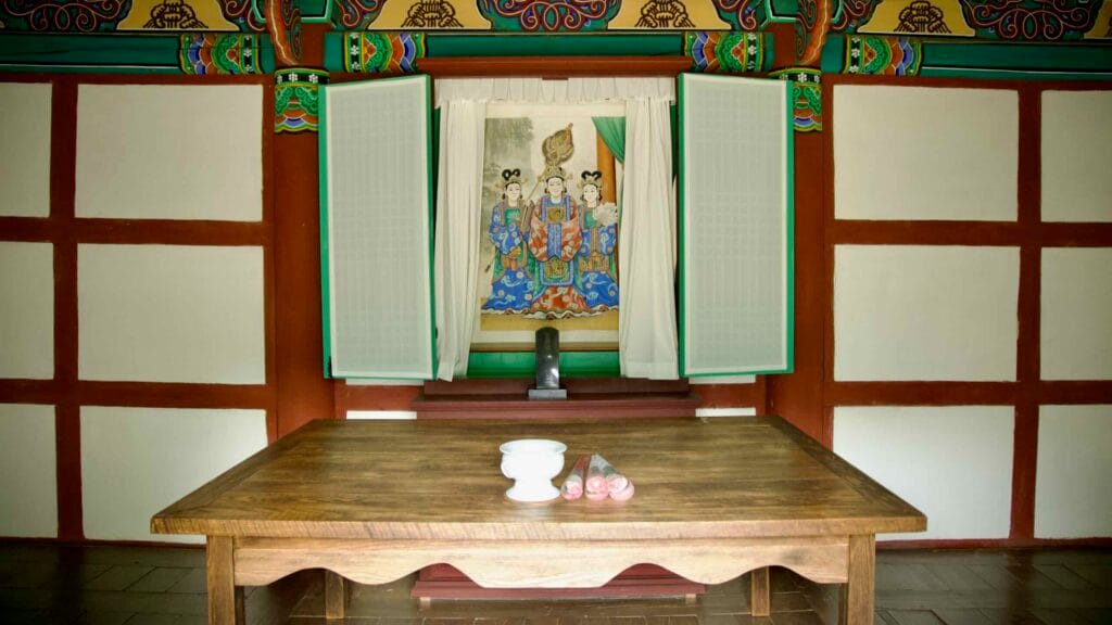 Inside Gungnyeosa Shrine's main hall, a wooden altar holds offerings beneath a painting of three royal women.