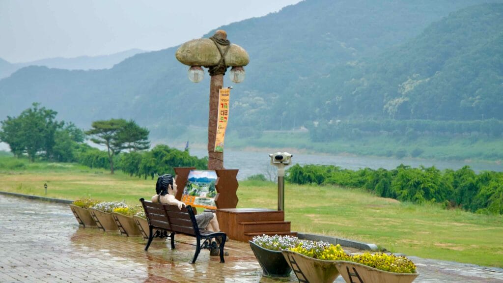 A sculpture of a prehistoric figure sits on a bench facing the Geum River at the Seokjangni Museum grounds.