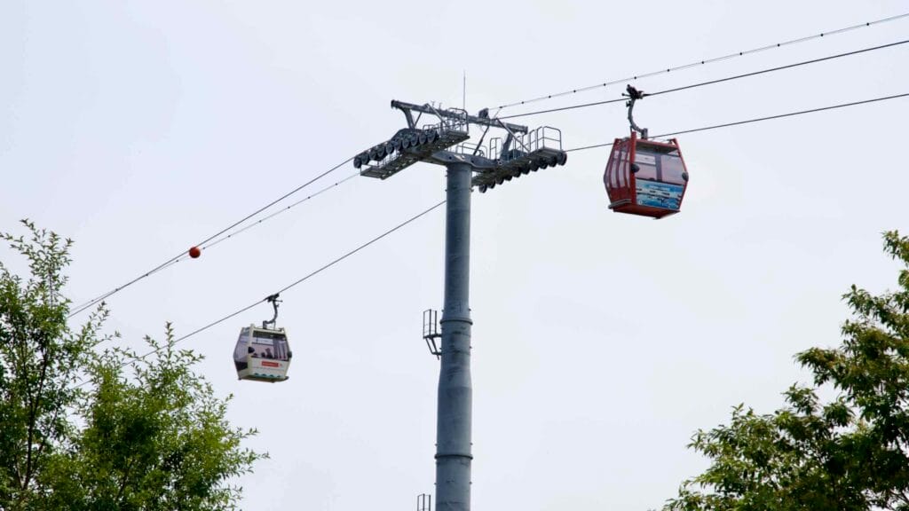 Two cable cars glide past a support tower along the Wangpicheon Cable Car route, offering passengers panoramic views above the trees and coastal hills.