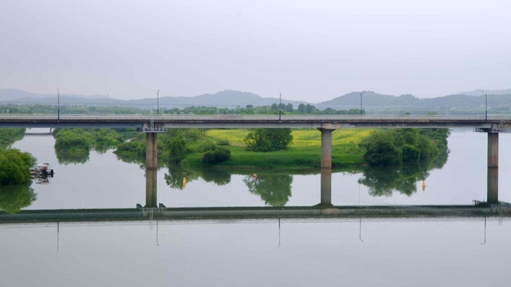 A road bridge stretches across the calm waters near Ipo Weir, reflecting the lush riverbanks and spring greenery of the South Han River basin.