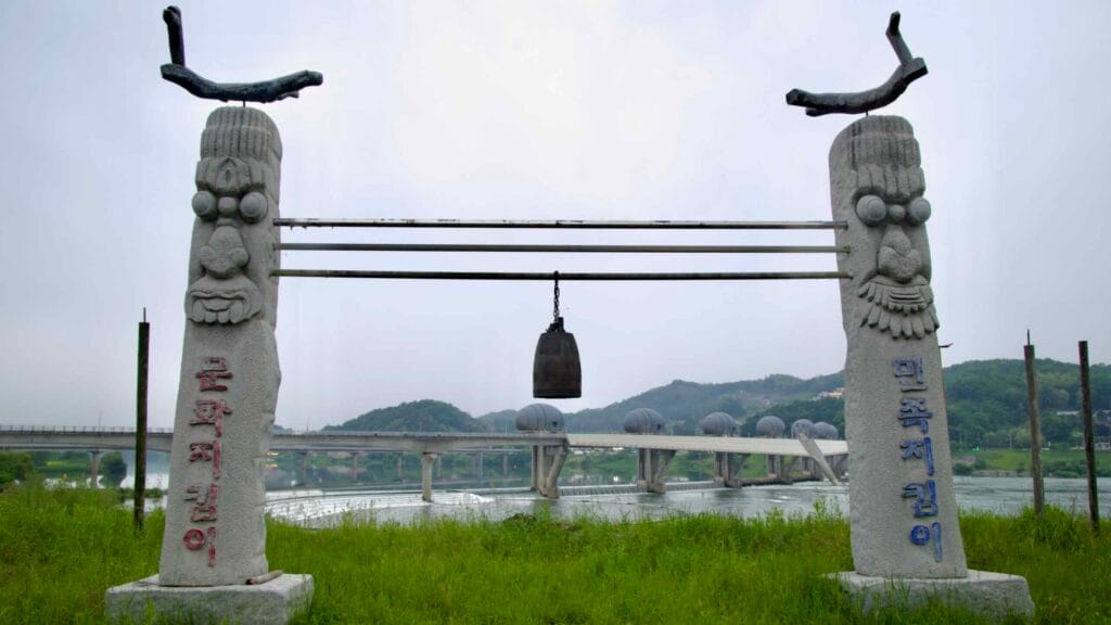A pair of jangseung totems stand with a hanging bell in front of Ipo Weir, blending Korean cultural heritage with modern water management infrastructure.