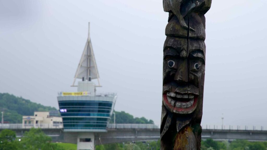 A carved wooden totem stands in sharp relief before the sail-shaped Ipo Weir Observatory, blending Korean folk heritage with modern riverfront design.