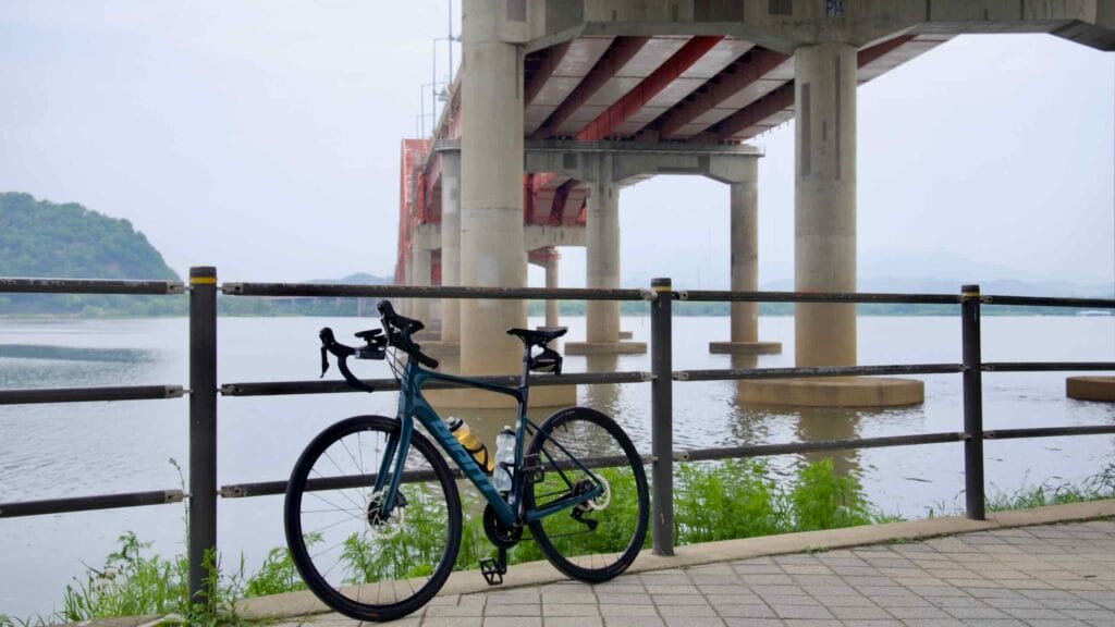 A road bicycle rests along the Han River beneath the towering concrete pillars of Banghwa Bridge, a quiet moment on the trail where infrastructure meets nature.