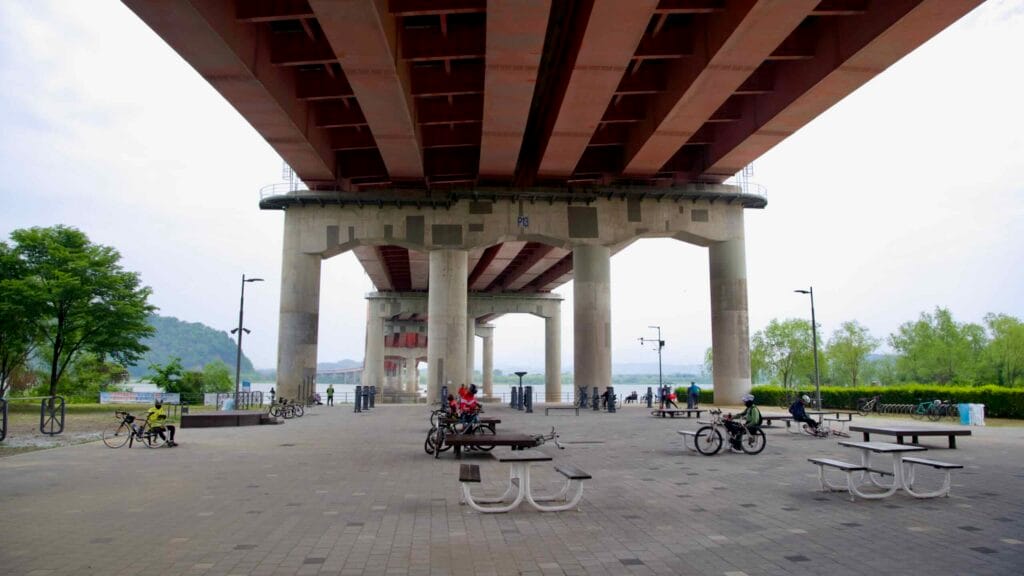 Cyclists take a break under Banghwa Bridge at a paved riverside plaza along the Han River, equipped with benches, tables, and bike racks sheltered beneath the overpass.
