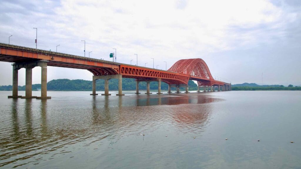 A wide-angle view of Banghwa Bridge shows its full red steel span arching over the Han River, connecting the western edge of Seoul to Incheon amid calm waters and cloudy skies.