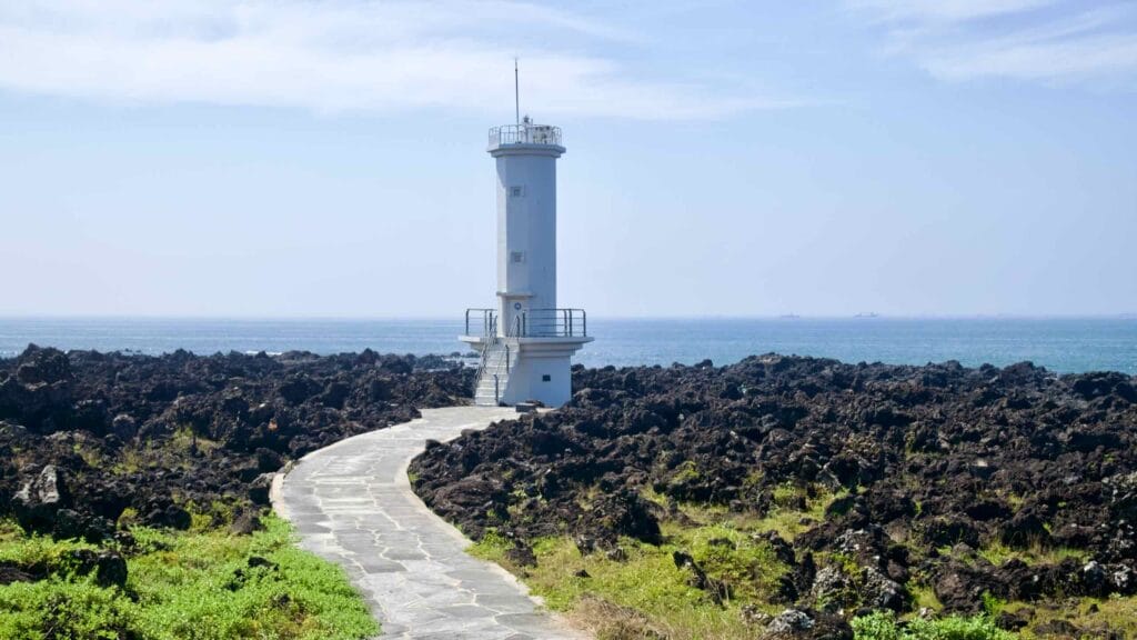 A stone footpath crosses jagged basalt to a small lighthouse on Jeju’s south coast near Sinheung-ri.