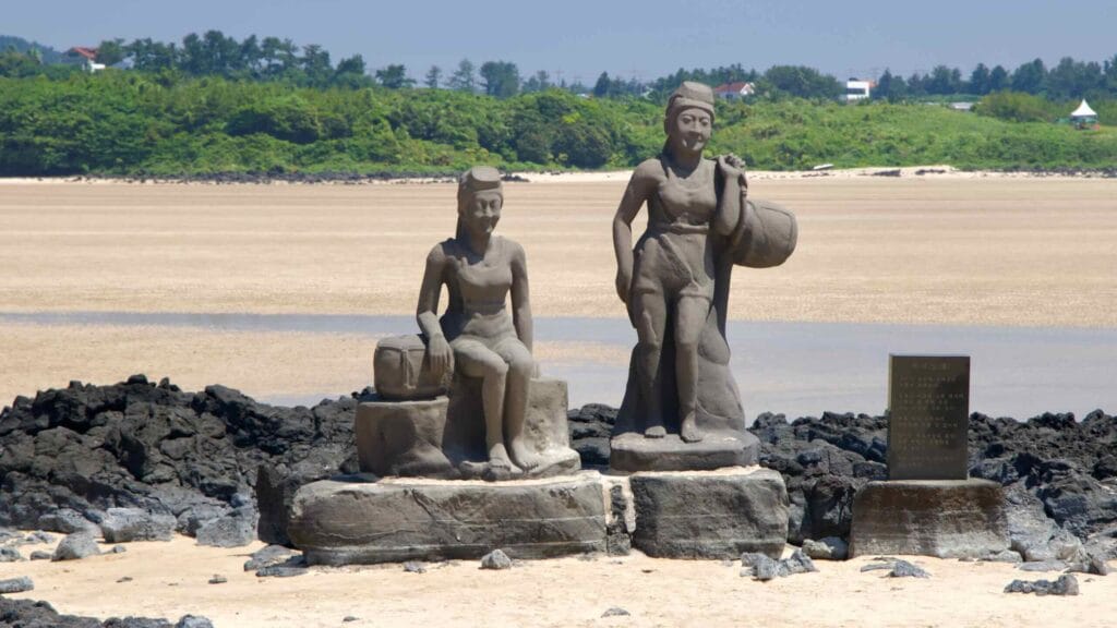 Twin haenyeo statues stand on basalt pedestals at Pyoseon Beach during low tide with sand and headlands behind.