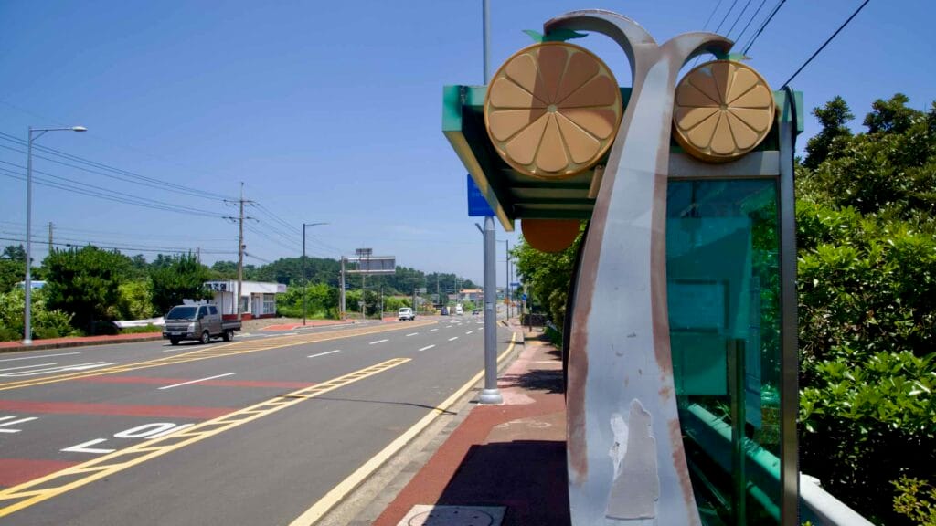 A citrus‑themed bus stop overlooks Iljudong Road with cars, power lines, and a bike lane.
